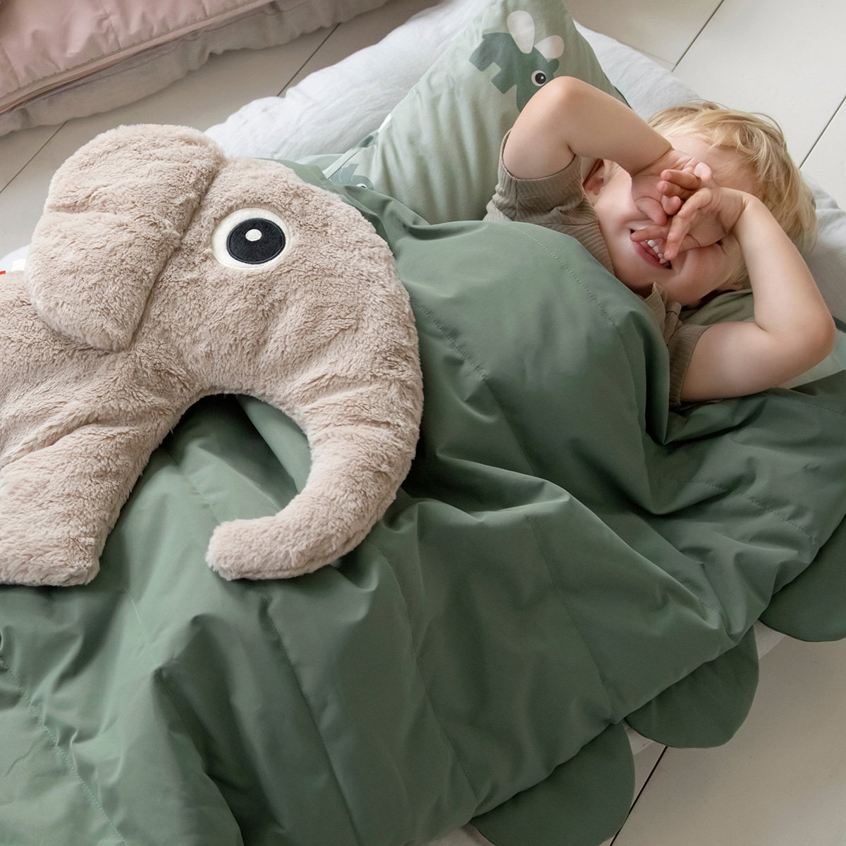 gosedjur elefant beige liggande på grön bädd intill ett barn som täcker ansiktet