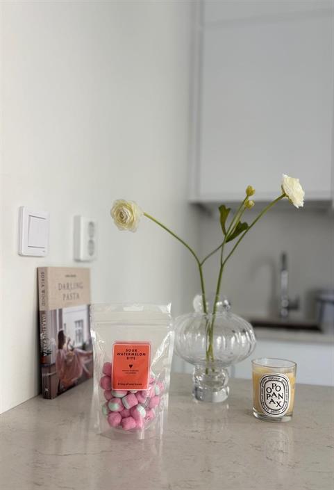 påse med sura vattenmelonbitar på köksbänk framför en glasvas med vita blommor och ett doftljus