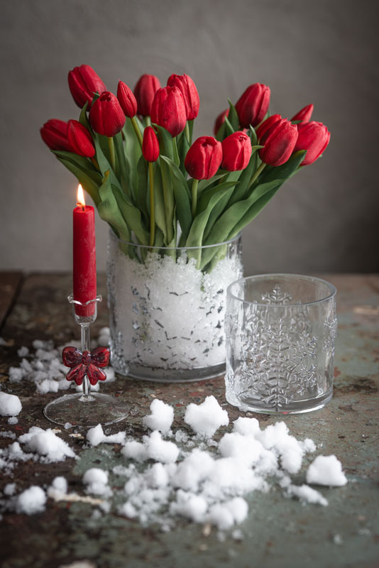 ljusstake rosett glas med brinnande rött kronljus framför vas med röda tulpaner och snö på bordet