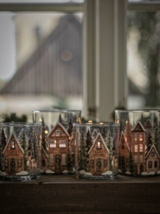 majas lykta rustic cottage i glas med vintermotiv av små stugor och granar, flera lyktor placerade på en fönsterbräda med varmt ljus inuti