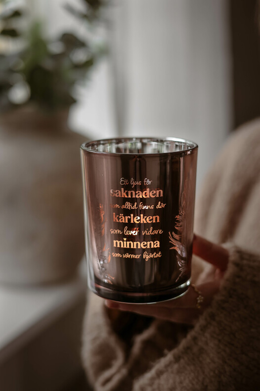 majas lykta saknaden i rökfärgat glas med upplyst ljus och gyllene text 'saknaden, kärleken, minnena', hålls i en hand framför en suddig inomhusbakgrund