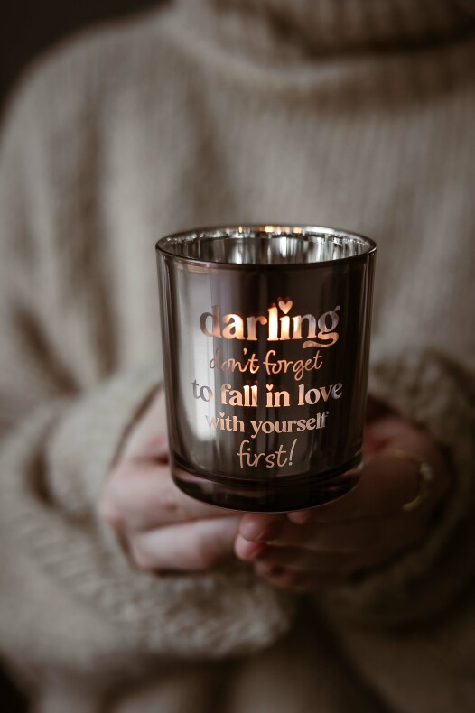 majas lykta darling i mörkt glas med guldfärgad text 'darling don't forget to fall in love with yourself first' hålls i två händer framför en beige stickad tröja
