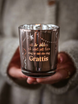 Majas lykta Grattis hålls i två händer, blank roséguldig glaslykta med tryckt text 'Grattis' och dekorativa mönster