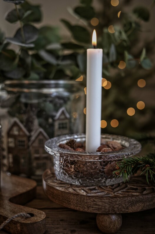 ljusstake i glas med ett vitt tänd kronljus, fylld med nötter och placerad på ett runt träunderlägg med juldekorationer