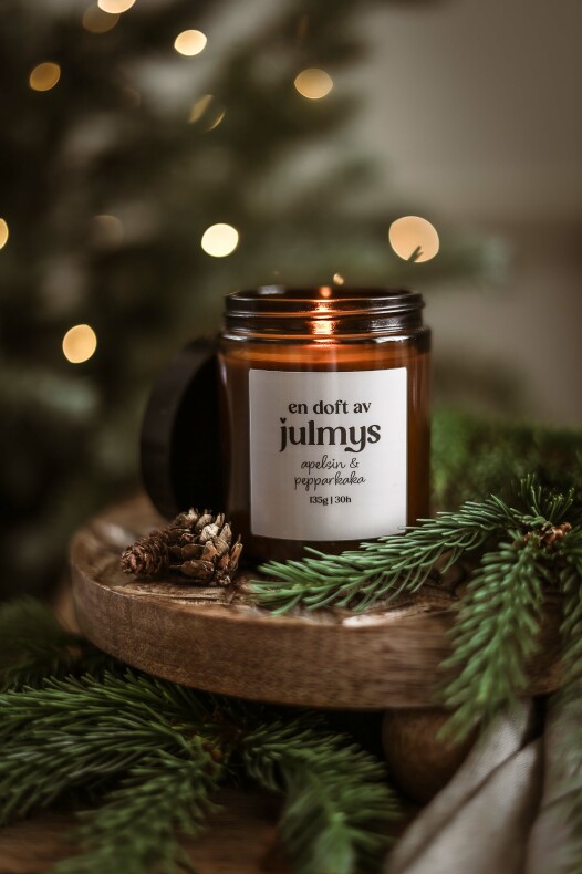 doftljus julmys i brunt glas med etiketten "en doft av julmys" på en träbricka omgiven av granris och kottar, varma ljuspunkter i bakgrunden