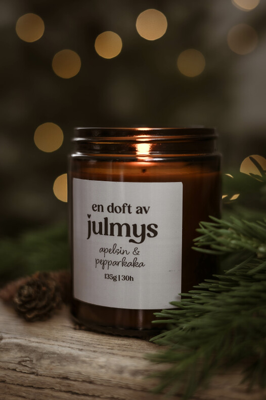 tätt foto på ett tänt doftljus julmys i en brun glasburk med etikett 'en doft av julmys apelsin & pepparkaka' på en träyta, omgivet av granris och kottar med varma bokeh-ljus i bakgrunden.