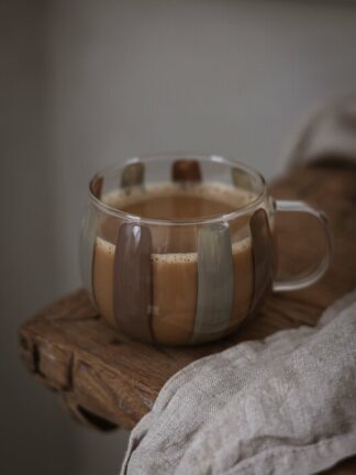 Majas glasmugg med breda vertikala ränder i brunt och grått, fylld med kaffe och placerad på en rustik träbräda.