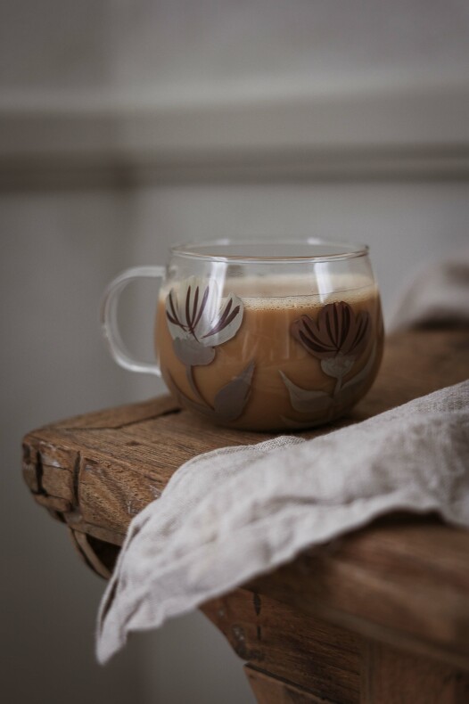 majas glasmugg blomia med bruna och vita blommotiv, fylld med kaffe på en rustik träbänk bredvid en linneservett