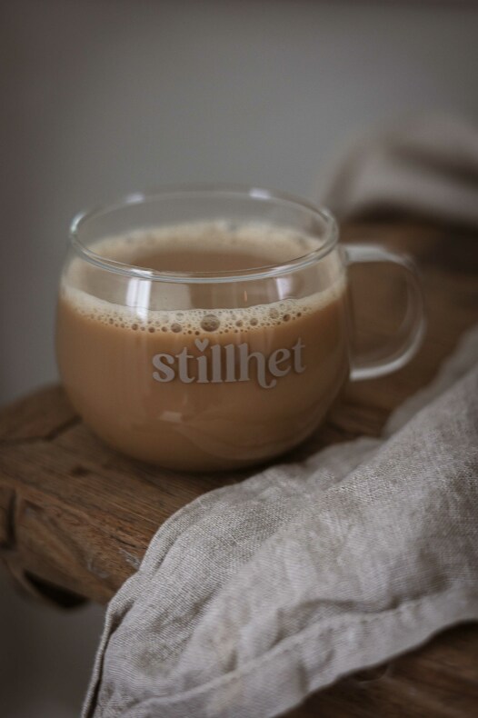 majas glasmugg stillhet i klart glas, fylld med ljusbrunt kaffe, placerad på ett träunderlägg intill en beige linnehandduk