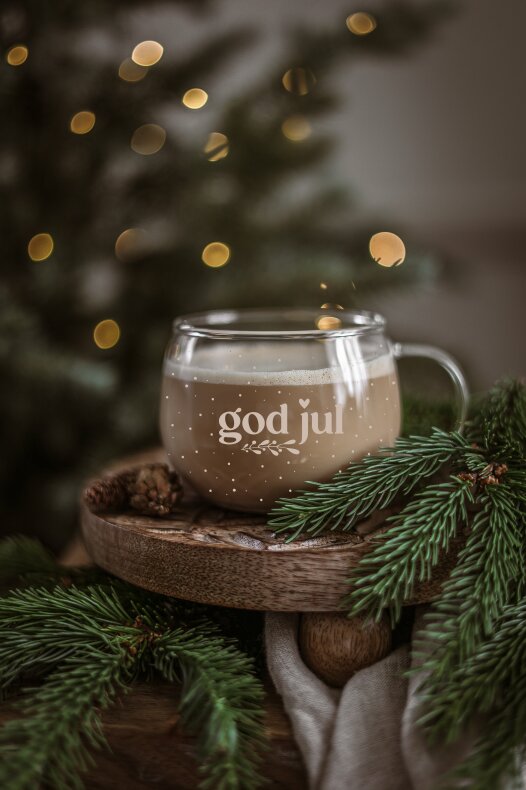 närbild på en glasmugg god jul fylld med varm dryck, placerad på en träplatta omgiven av granris och oskarpa julgransljus.