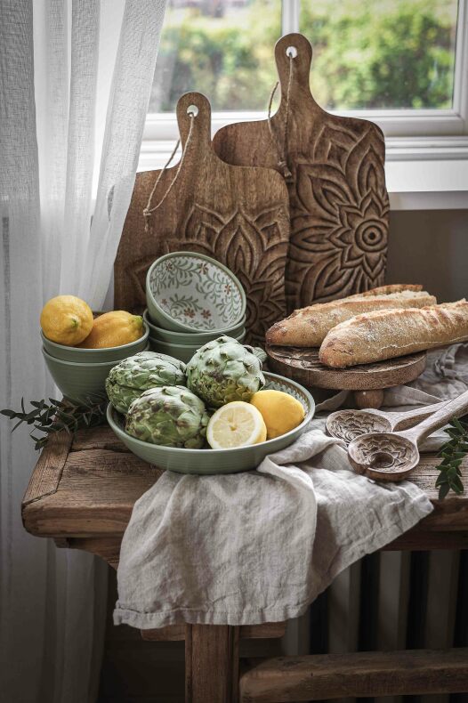 tre träskärbrädor med snidat blommotiv, en dekorerad skärbräda i mitten står på ett rustikt bord med skålar, kronärtskockor, citronhalvor, träskedar och baguetter vid ett fönster.