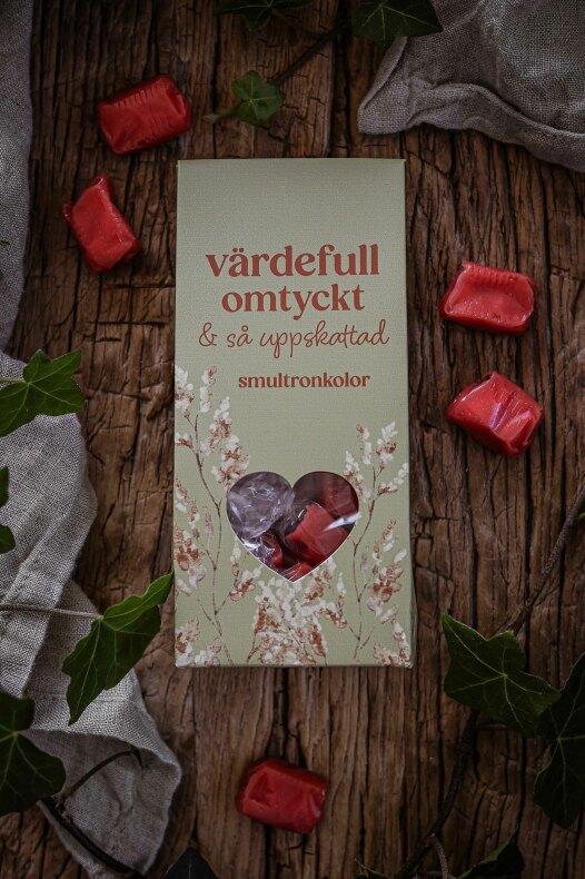 majas smultronkolor - grön presentförpackning med hjärtformat fönster som visar inlindade röda kolor, placerad på träbakgrund med gröna blad och lösa röda kolor runtom.