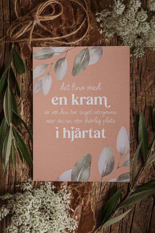 majas vykort en kram — rosa kort med vit text 'en kram' och bladillustrationer på träbakgrund, omgiven av gröna blad, vita blommor och ett snöre