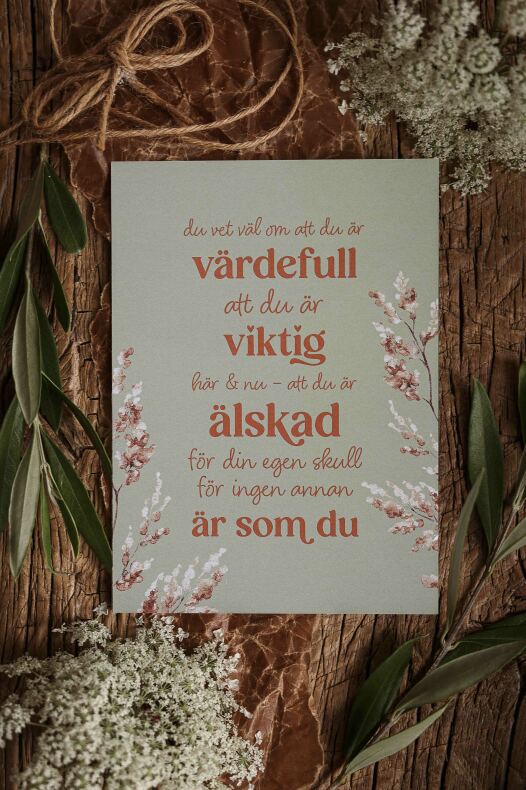 majas vykort med texten "du vet väl om att du är värdefull" i varm orange typografi på ljusgrön bakgrund, omgiven av blommor, gröna blad och ett snöre på träunderlag