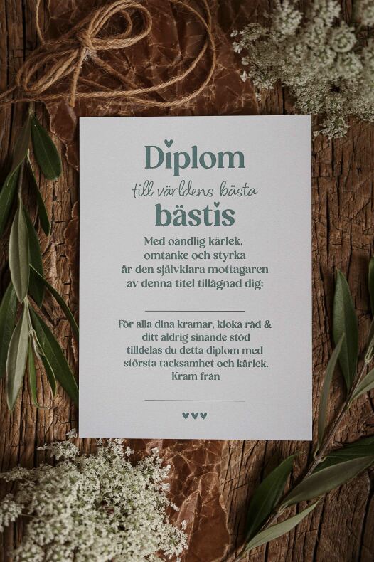 vitt kort med tryckt text 'diplom till världens bästa bästis' liggande på träbakgrund omgivet av gröna blad, vita blommor och ett snöre — diplom till bästis
