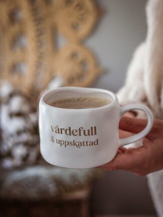 Vit keramikmugg med brun text ”värdefull & uppskattad”, fylld med kaffe och hållen i en hand.