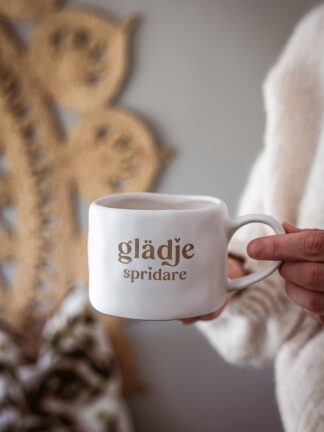 Vit keramikmugg med brun text ”glädje spridare”, hållen i en hand framför en rottingdekoration på väggen.