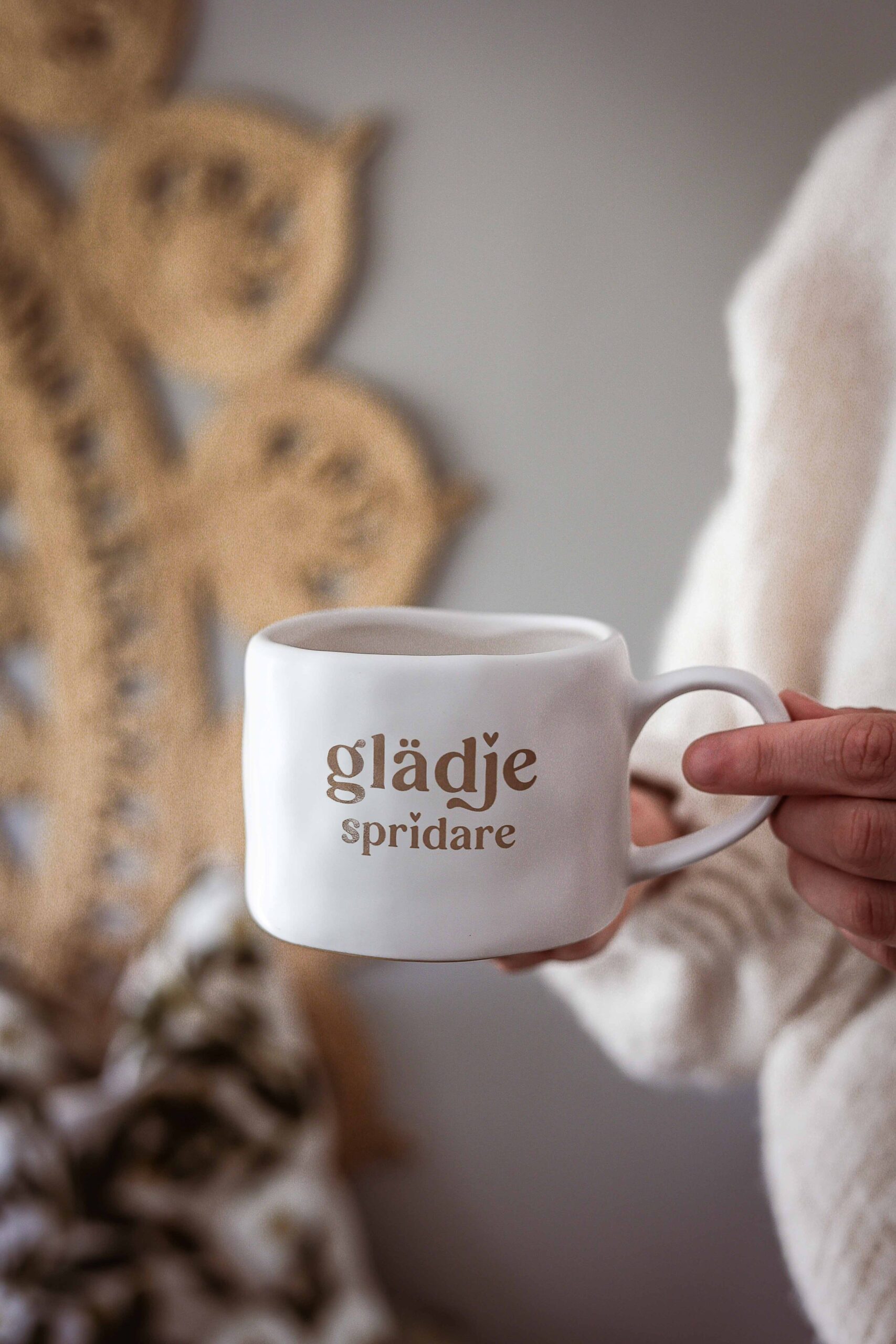 vit keramikmugg med brun text ”glädje spridare”, hållen i en hand framför en rottingdekoration på väggen.