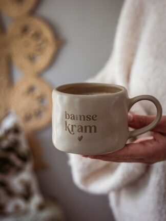 Sandfärgad keramikmugg med brun text ”bamse kram” och ett hjärta under, fylld med kaffe och hållen i en hand.