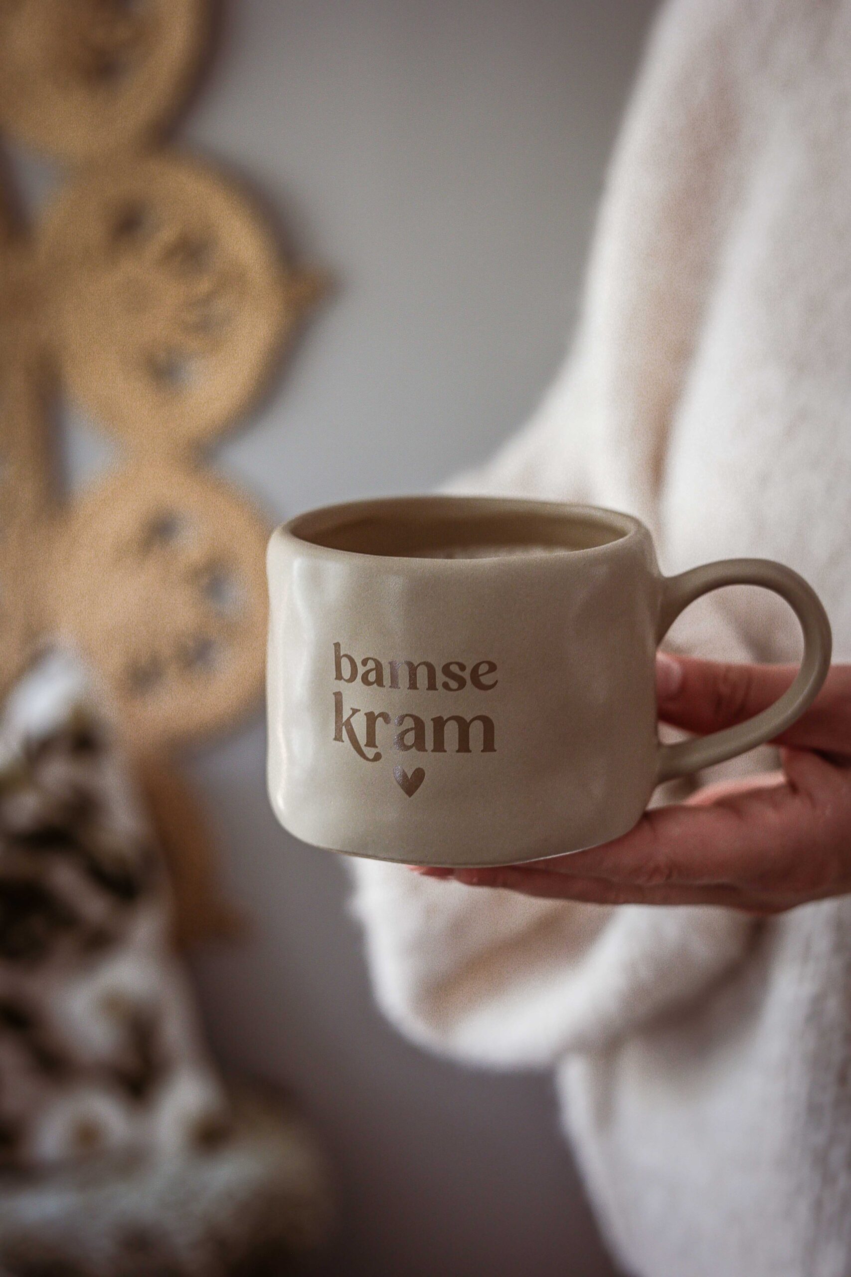 sandfärgad keramikmugg med brun text ”bamse kram” och ett hjärta under, fylld med kaffe och hållen i en hand.