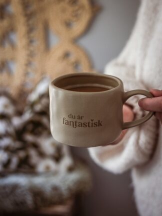 Beige keramikmugg med brun text ”du är fantastisk” och ett litet hjärta, fylld med kaffe och hållen i en hand.