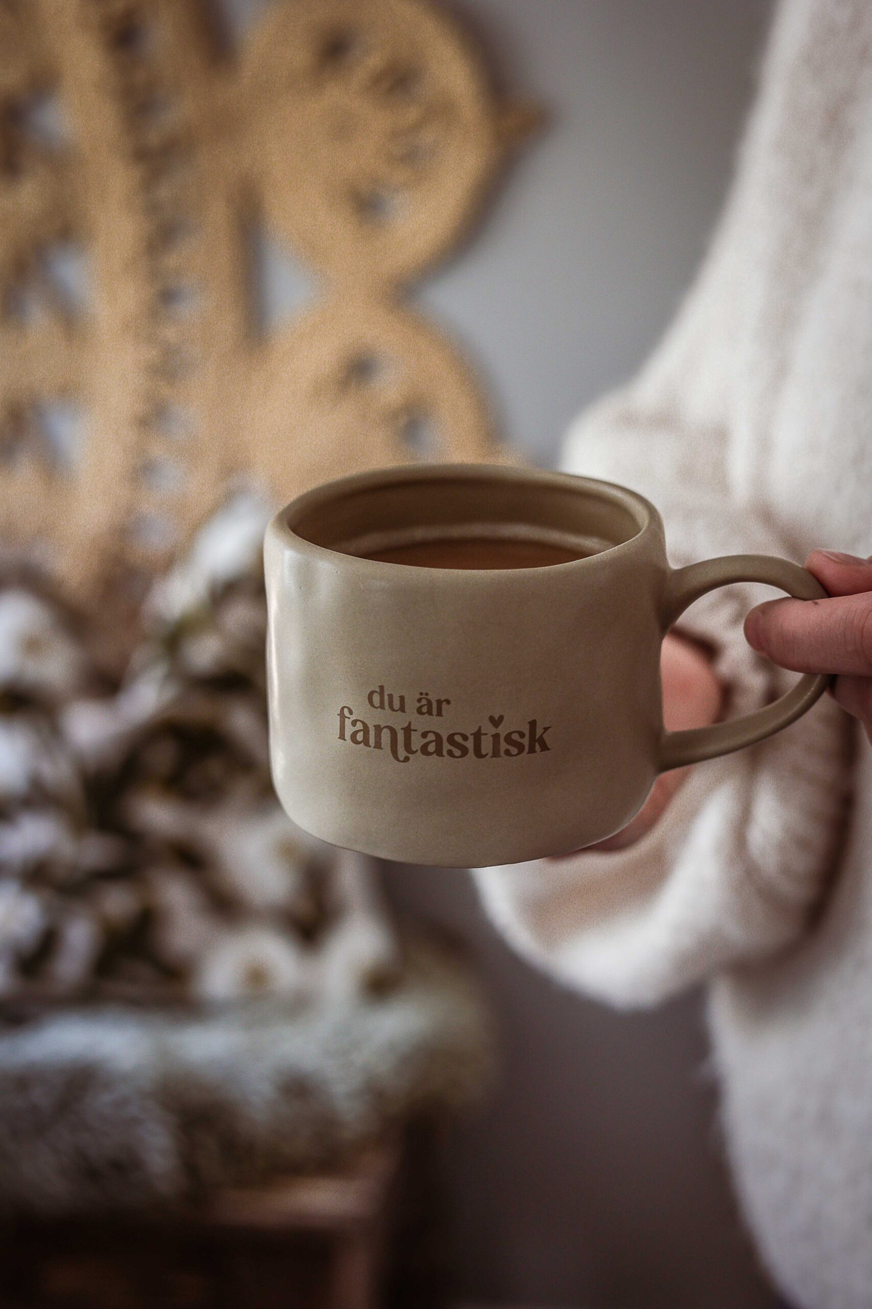 beige keramikmugg med brun text ”du är fantastisk” och ett litet hjärta, fylld med kaffe och hållen i en hand.