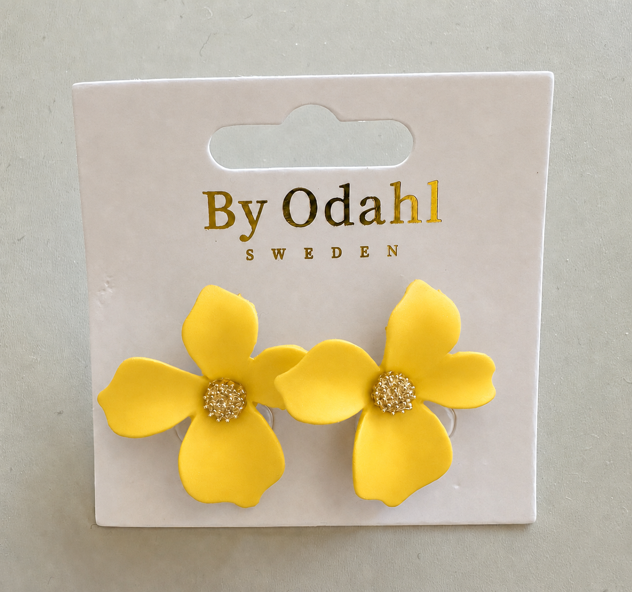 närbild på gul blomma örhängen, två gula blomformade örhängen på ett vitt upphängningskort med märket 'by odahl'.